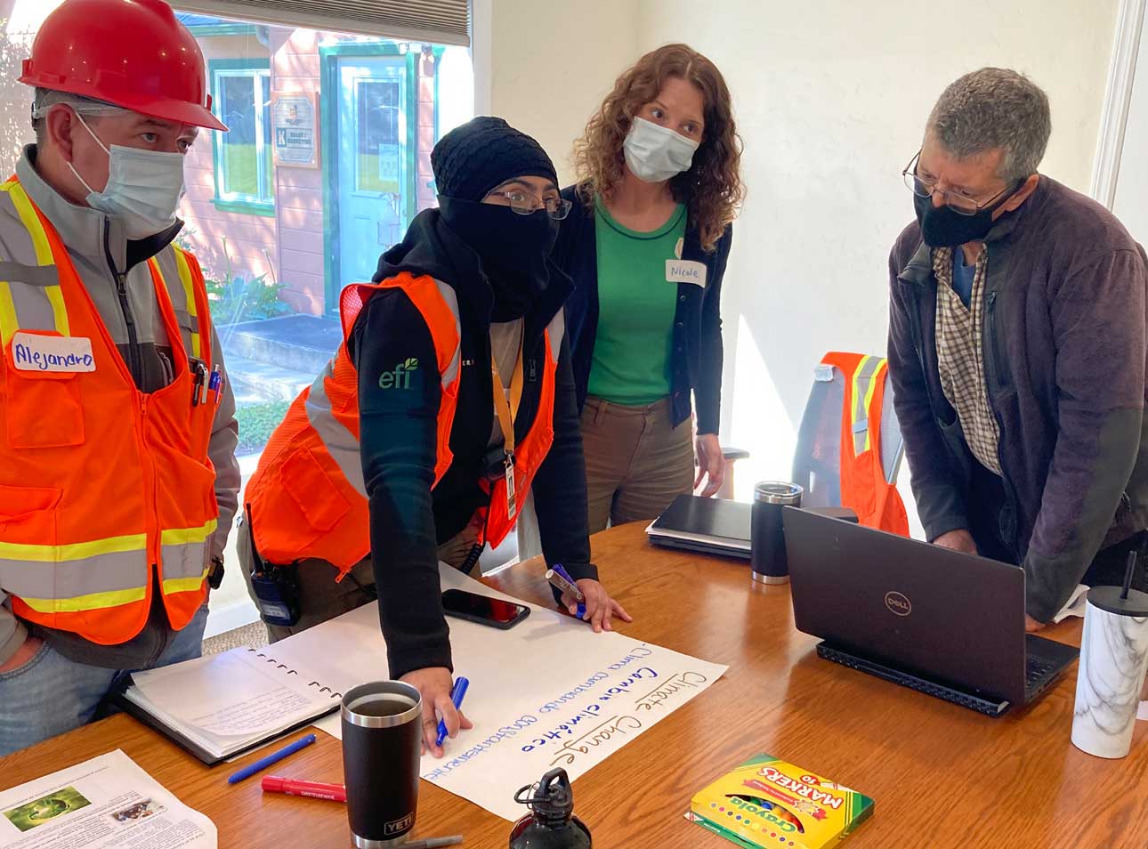 Two women and two men dressed in safety clothes gather around a desk with a laptop during Misionero training